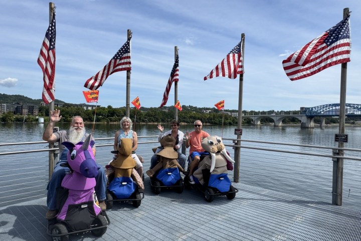 Four people on animal-themed scooters by a river, with American flags and a bridge in the background.