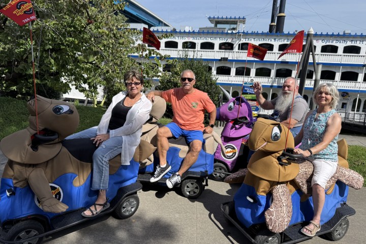 Four people ride motorized animal scooters near a riverboat on a sunny day.
