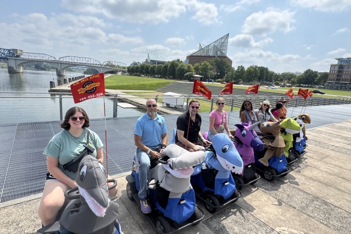 Group of people riding animal scooters near a river with a bridge and modern building in the background.