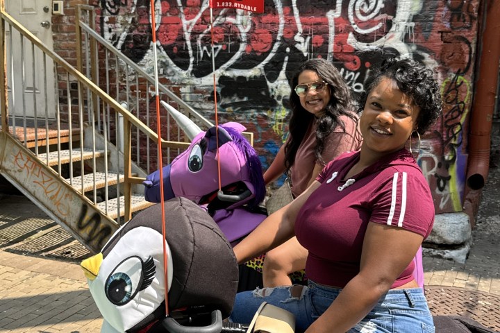 Two women sitting on decorated scooters in front of a graffiti-covered wall.
