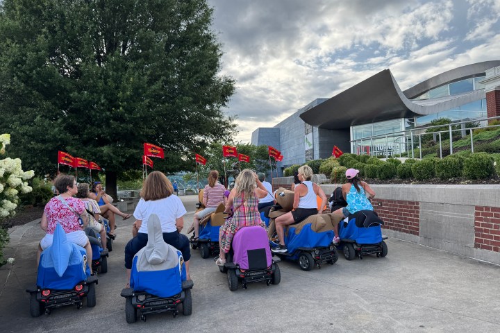 Group riding mobility scooters outside wavy-roofed building with red flags and tree nearby.