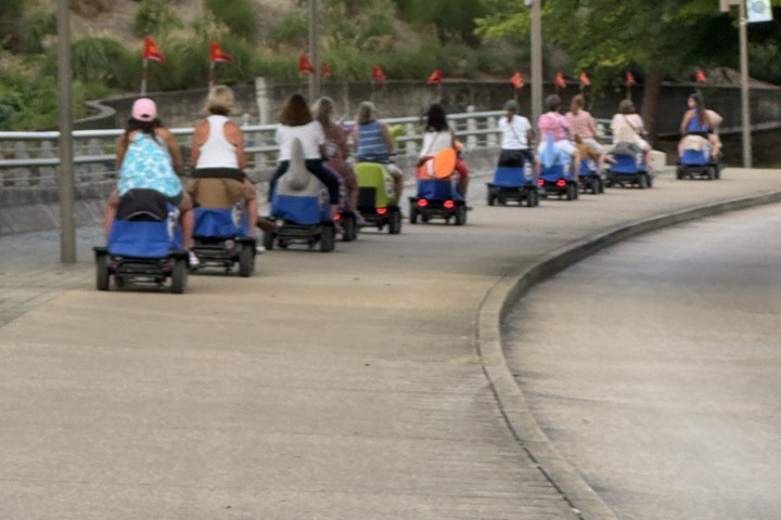 Group of people on motorized scooters riding on a paved path with greenery around.