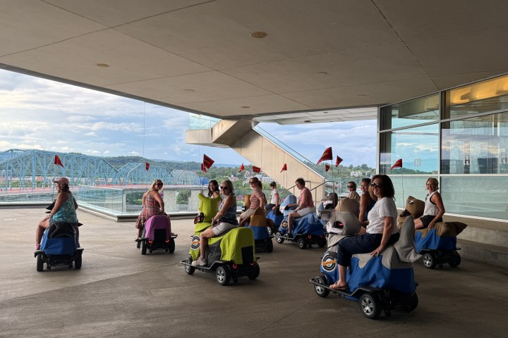 Group of people on motorized scooters under a building canopy near a bridge.