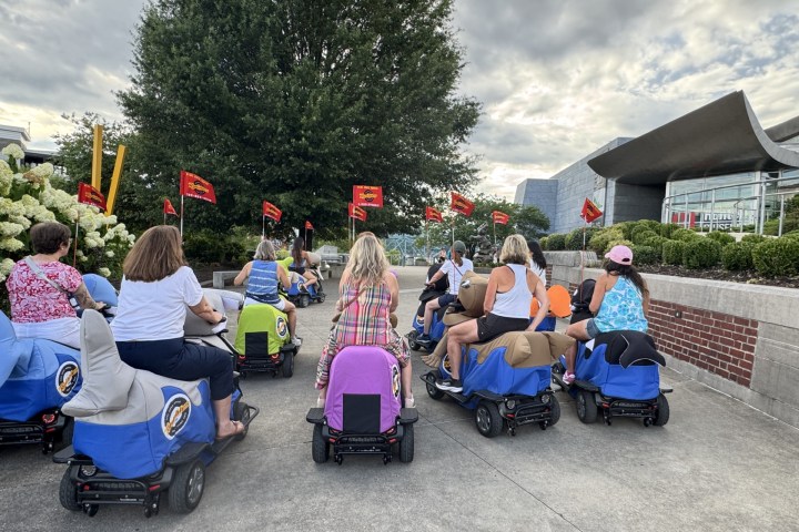 Group of people on mobility scooters with colorful covers, outdoors near modern building and trees.