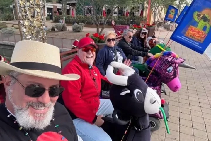 Group of people in festive attire riding mobility scooters shaped like animals on a city street.