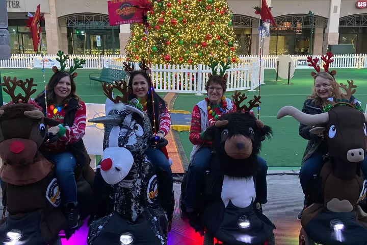 Four people on animal-themed scooters in front of a large decorated Christmas tree.