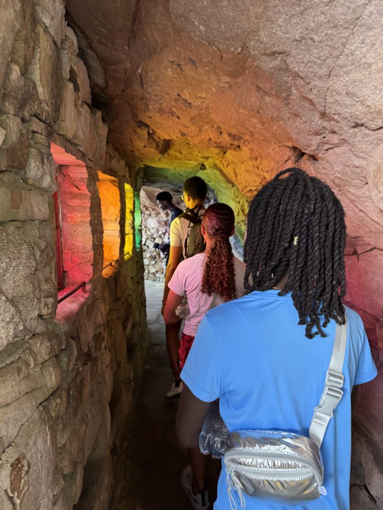People walking through a rocky tunnel illuminated by rainbow lights.