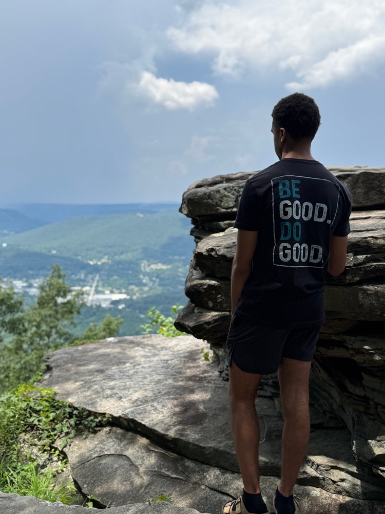 Person in shirt reading 'Be Good. Do Good.' stands on cliff overlooking hills.