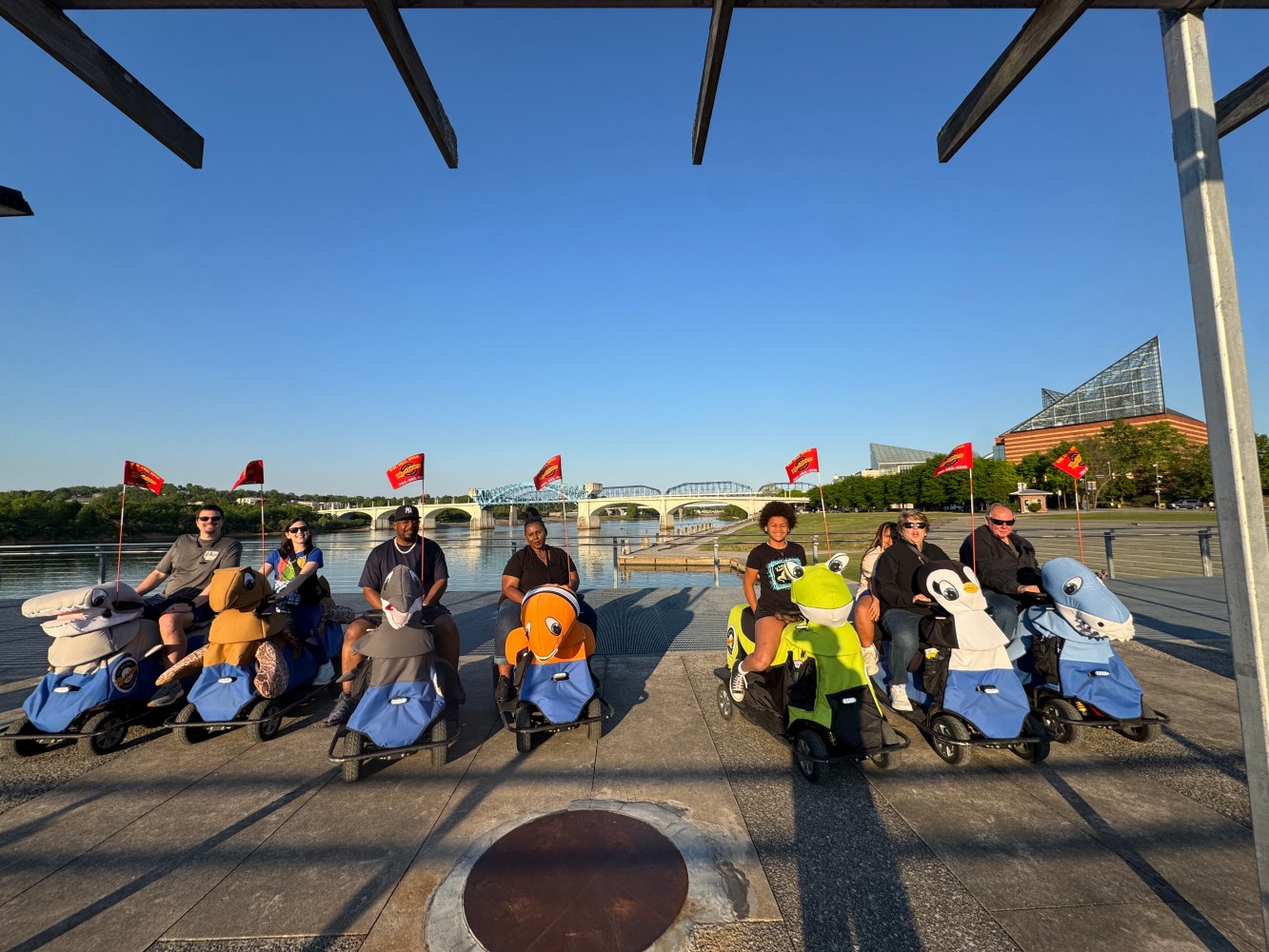 Group on animal scooters at a riverside park with bridge in background.