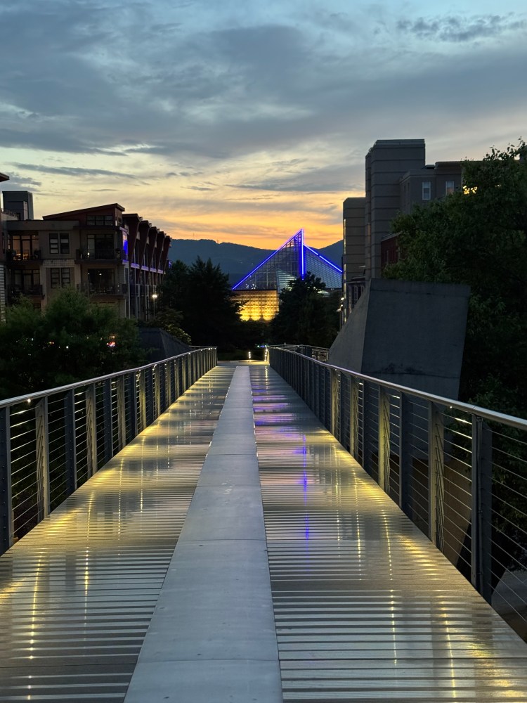 Illuminated bridge leading to a lit blue building under a cloudy sunset sky.