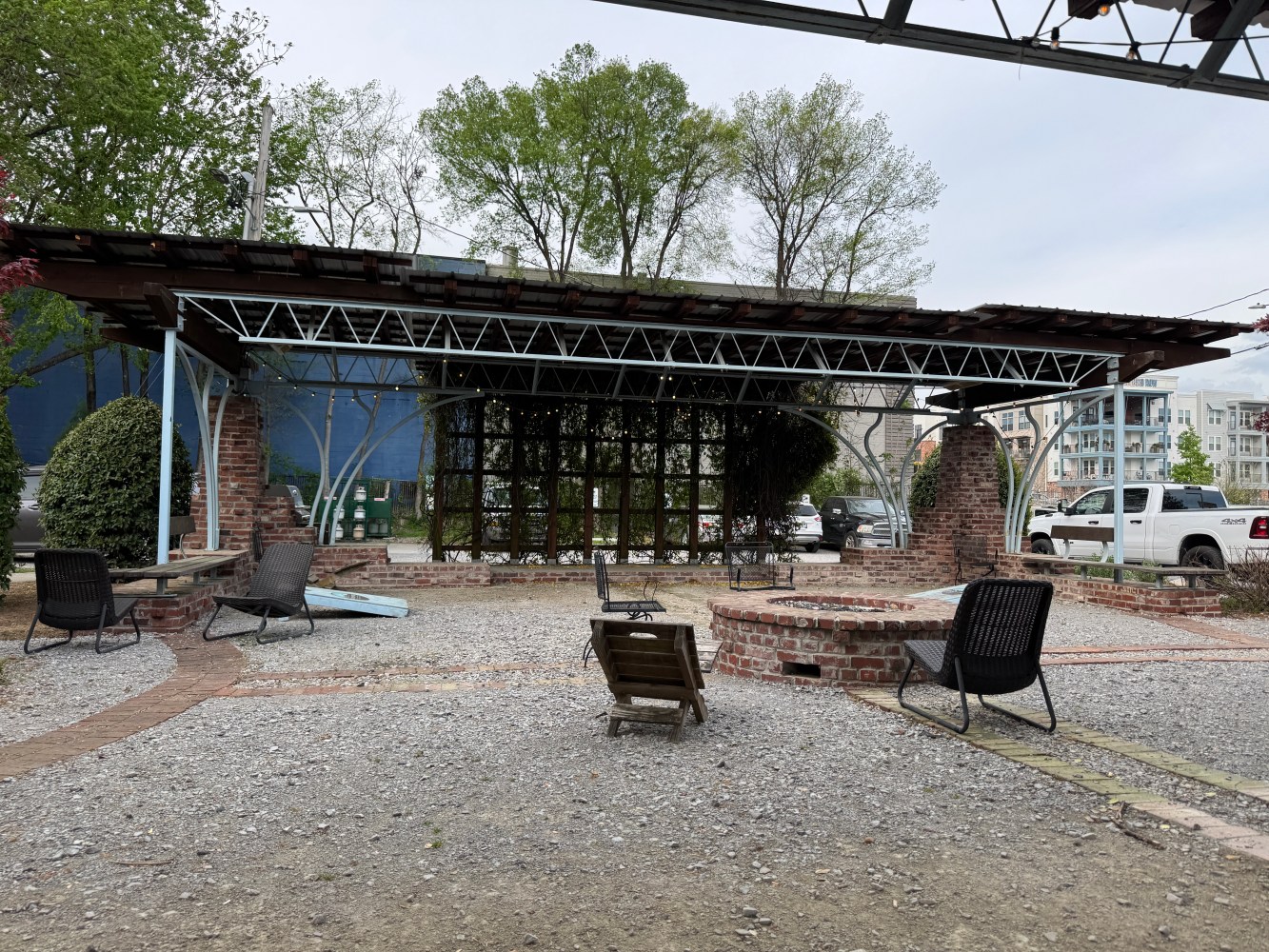 Outdoor seating area with metal structure and chairs on gravel surface, surrounded by trees.