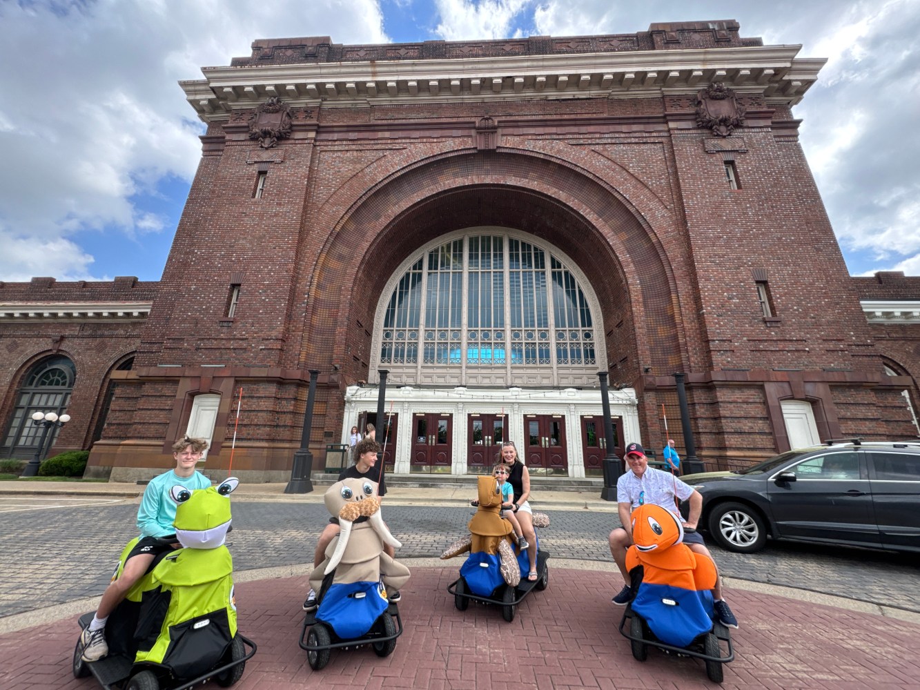 People riding animal scooters in front of a large brick building with an arched window.